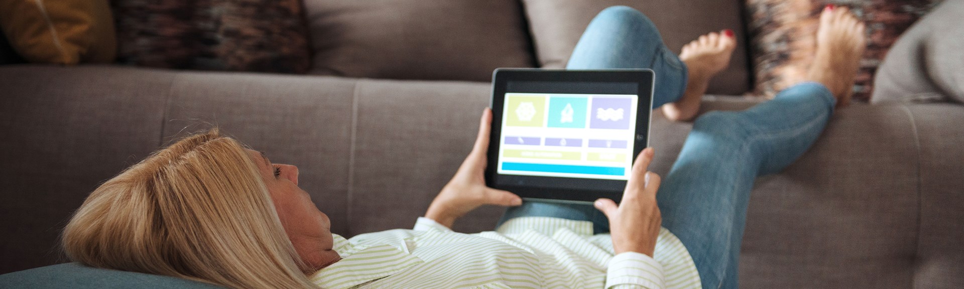 Smarthome Wärmepaket Eine Frau liegt auf der Couch, mit den Füßen auf der Lehne und schaut sich auf ihrem Tablet etwas an.